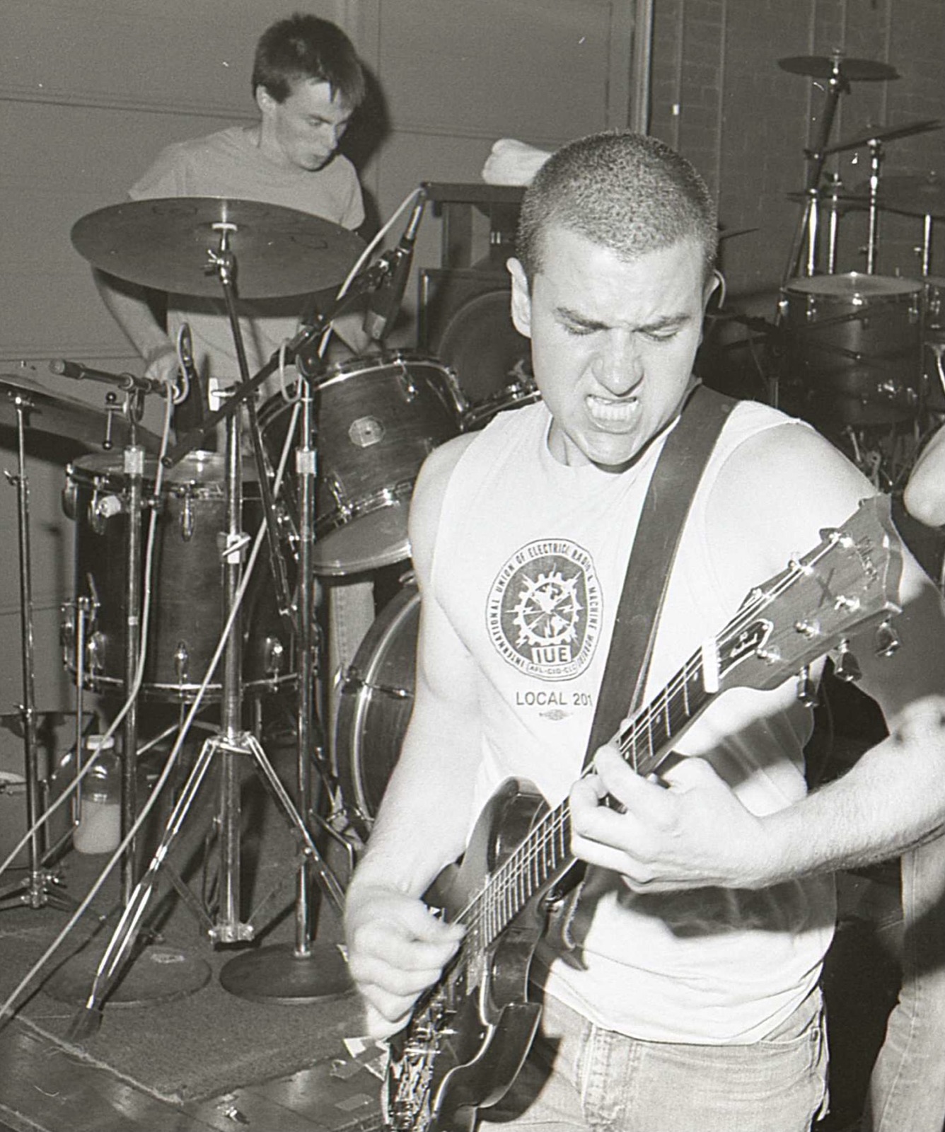 Guitarist Al Barile grimacing as he digs into his Gibson SG, wearing an IUE Local 201 union shirt, with the drummer playing behind him.