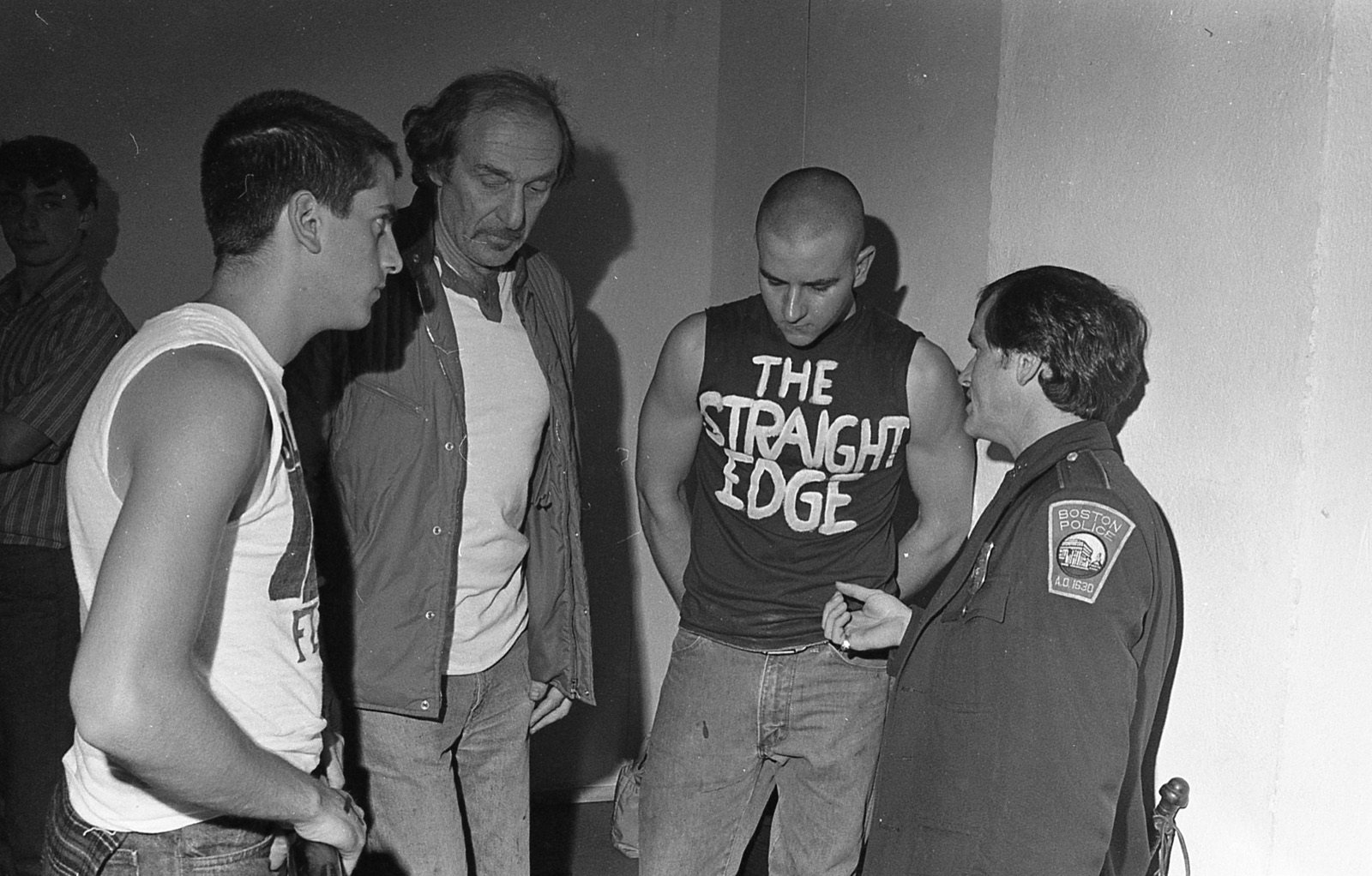 Three young men, one in a shirt hand-painted with The Straight Edge, listening to a Boston Police officer backstage.