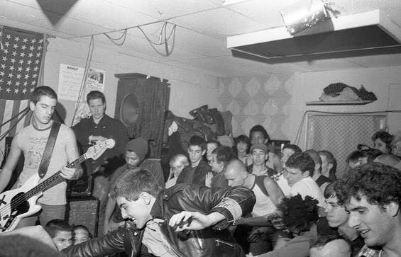 SSD playing a packed low-ceilinged room with the bassist at the left and dancers spilling into the band beneath an American flag.