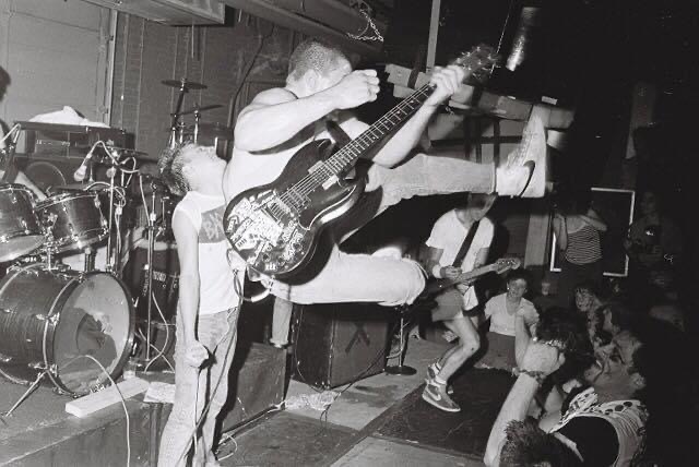 An SSD band member leaps through the air with his bass mid-song while the singer holds the microphone and the crowd watches from beside the stage.