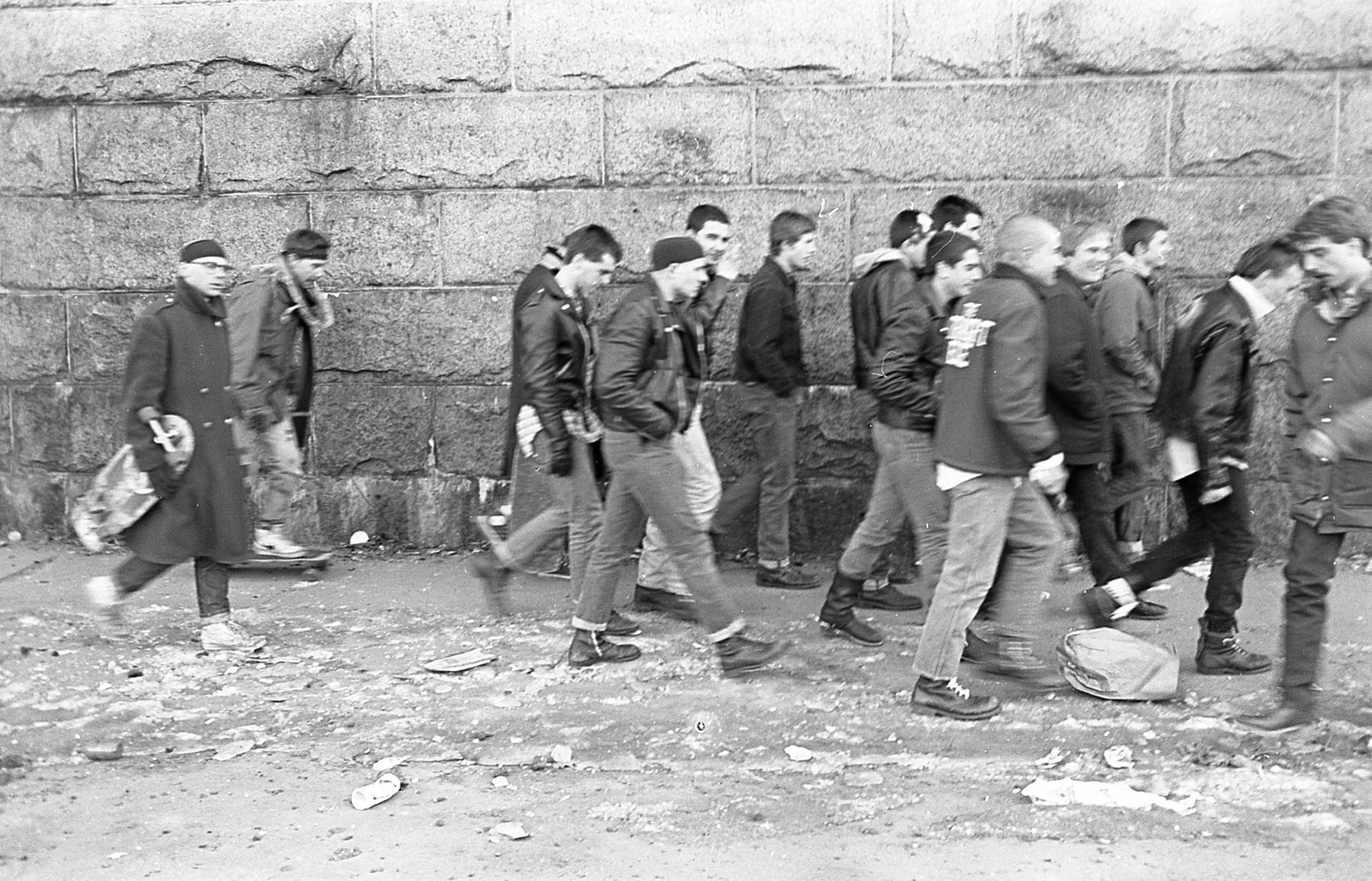 The Boston Crew walking in a long line beneath a granite wall, one of them rolling along on a skateboard.