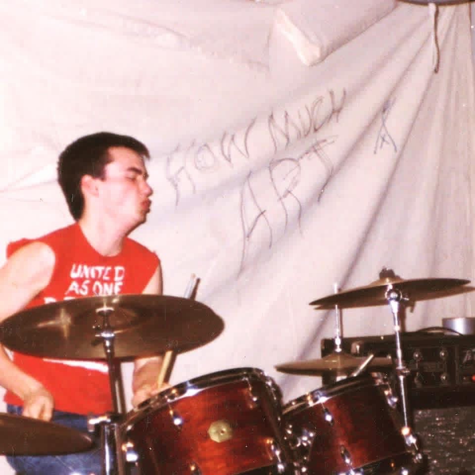 SSD's drummer in a red United As One band shirt playing his kit, with How Much Art written on the sheet hanging behind him.