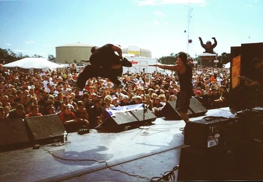 A member of GAGE flies through the air above an outdoor festival stage while the singer points and a huge crowd looks on.