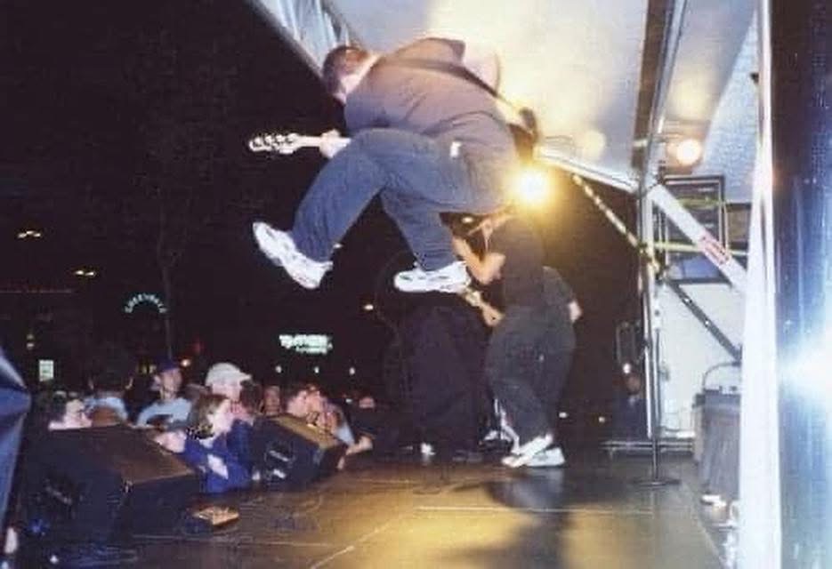 A GAGE guitarist leaps high above the stage at a nighttime outdoor show, guitar in hand, with the crowd below.
