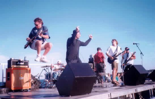 GAGE playing an outdoor stage under a clear blue sky, the guitarist mid-leap and the singer with a fist in the air.