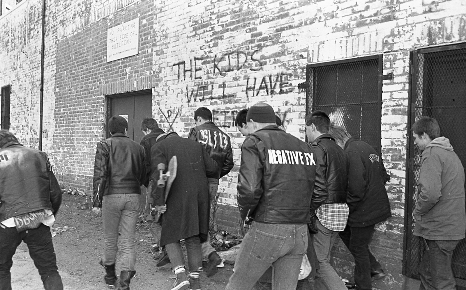 The Boston Crew walking past a brick wall spray-painted with The Kids Will Have Their Say.