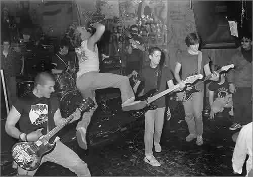 SS Decontrol playing live on a packed club stage, the singer leaping into the air mid-song beside the guitarists and bassist.