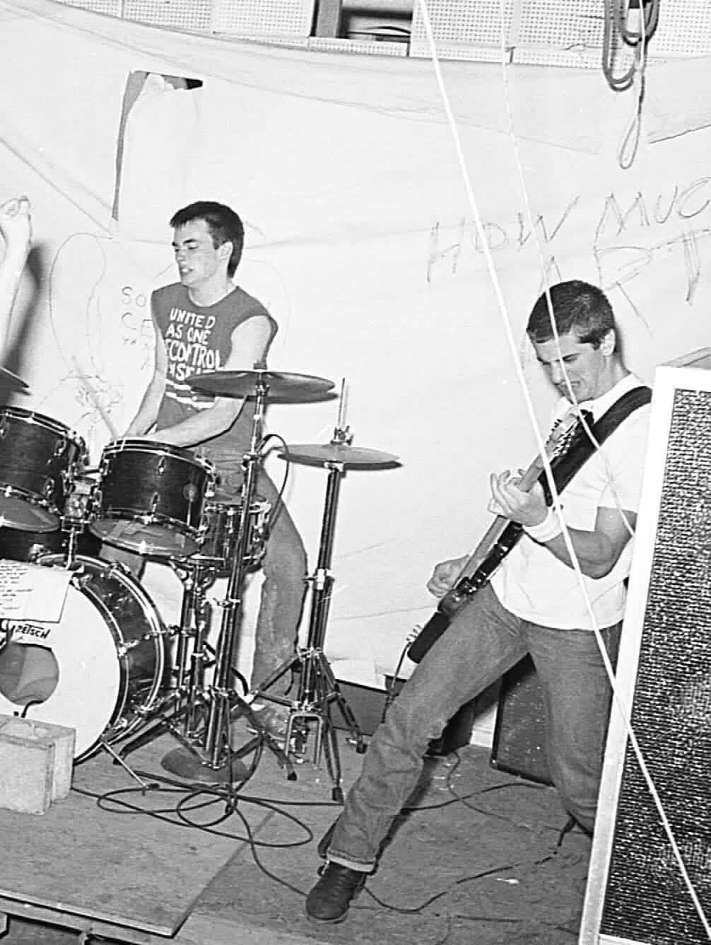 SSD's drummer, in a band shirt reading United As One, and a guitarist playing on stage in front of a hand-painted How Much Art backdrop.