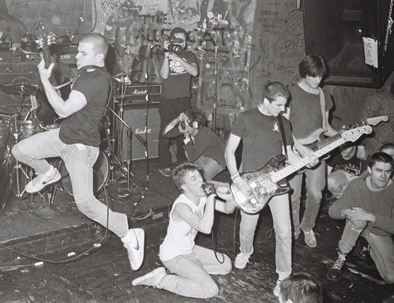 SSD playing live in a graffiti-covered club, the singer kneeling into the microphone while a guitarist jumps and the crowd crouches at the edge of the stage.